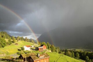 En dobbel regnbue spenner seg over et landlig landskap med grønne åser, spredte hus, trær og fjell i det fjerne under en delvis overskyet himmel.