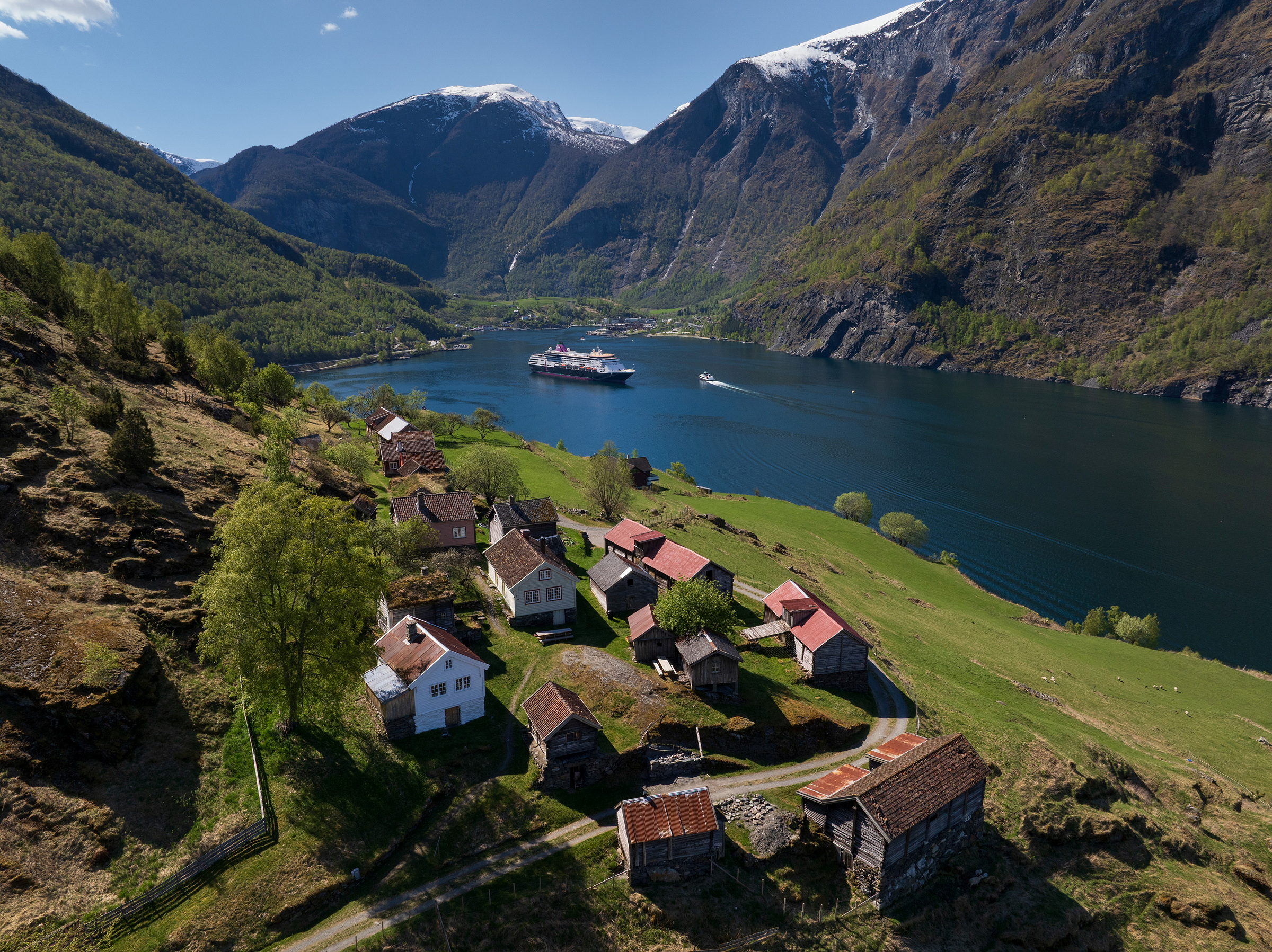 En liten landsby med trehus ligger på en grønn åsside over en fjord, med et cruiseskip og en båt på vannet, omgitt av bratte, skogkledde fjell under en klar himmel.
