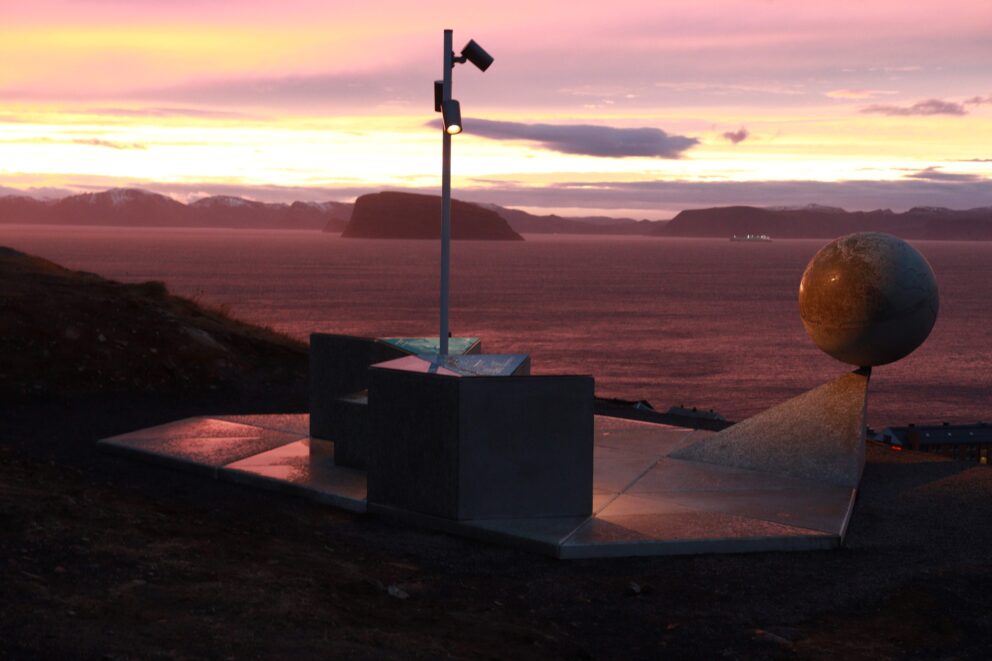 Verdsarvutstillinga på plass i Hammerfest 8 Et globusmonument og en enkelt lyktestolpe på en klippe med utsikt over en bukt ved solnedgang.