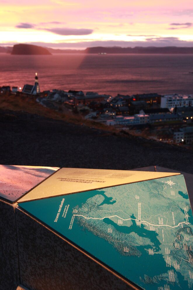 Verdsarvutstillinga på plass i Hammerfest 6 En informasjonsplakett i metall med et kart over Struves geodetiske bue, med utsikt over en kystby og havnen ved solnedgang.