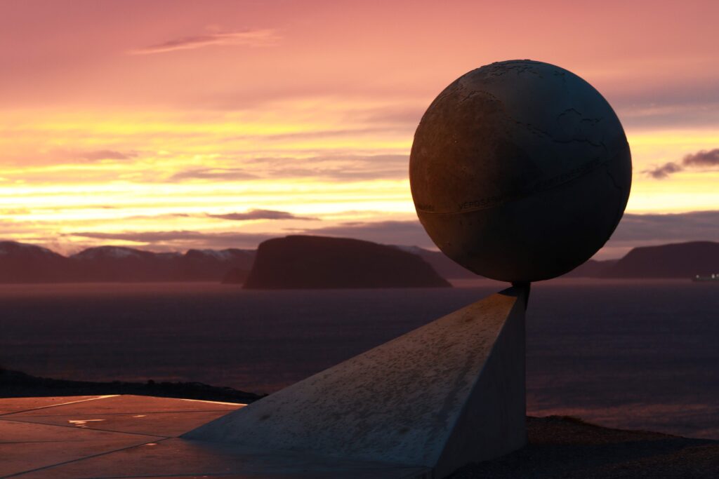 En globusskulptur i metall, montert på en trekantet sokkel, vender ut mot et mørkt hav og fjell i det fjerne under en fargerik solnedgang.