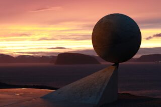 En globusskulptur i metall, montert på en trekantet sokkel, vender ut mot et mørkt hav og fjell i det fjerne under en fargerik solnedgang.