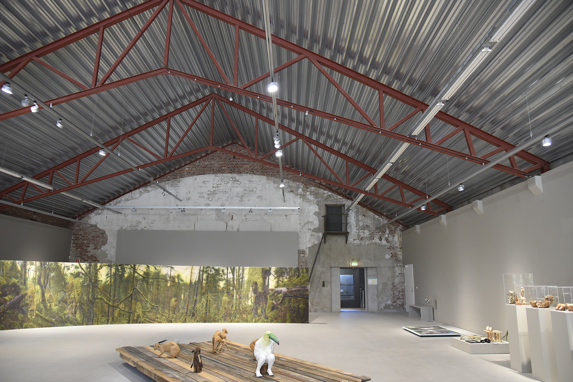 Et galleri for moderne kunst med høye tak, synlige bjelker og dyreskulpturer i blandede materialer utstilt på treplattformer i midten.