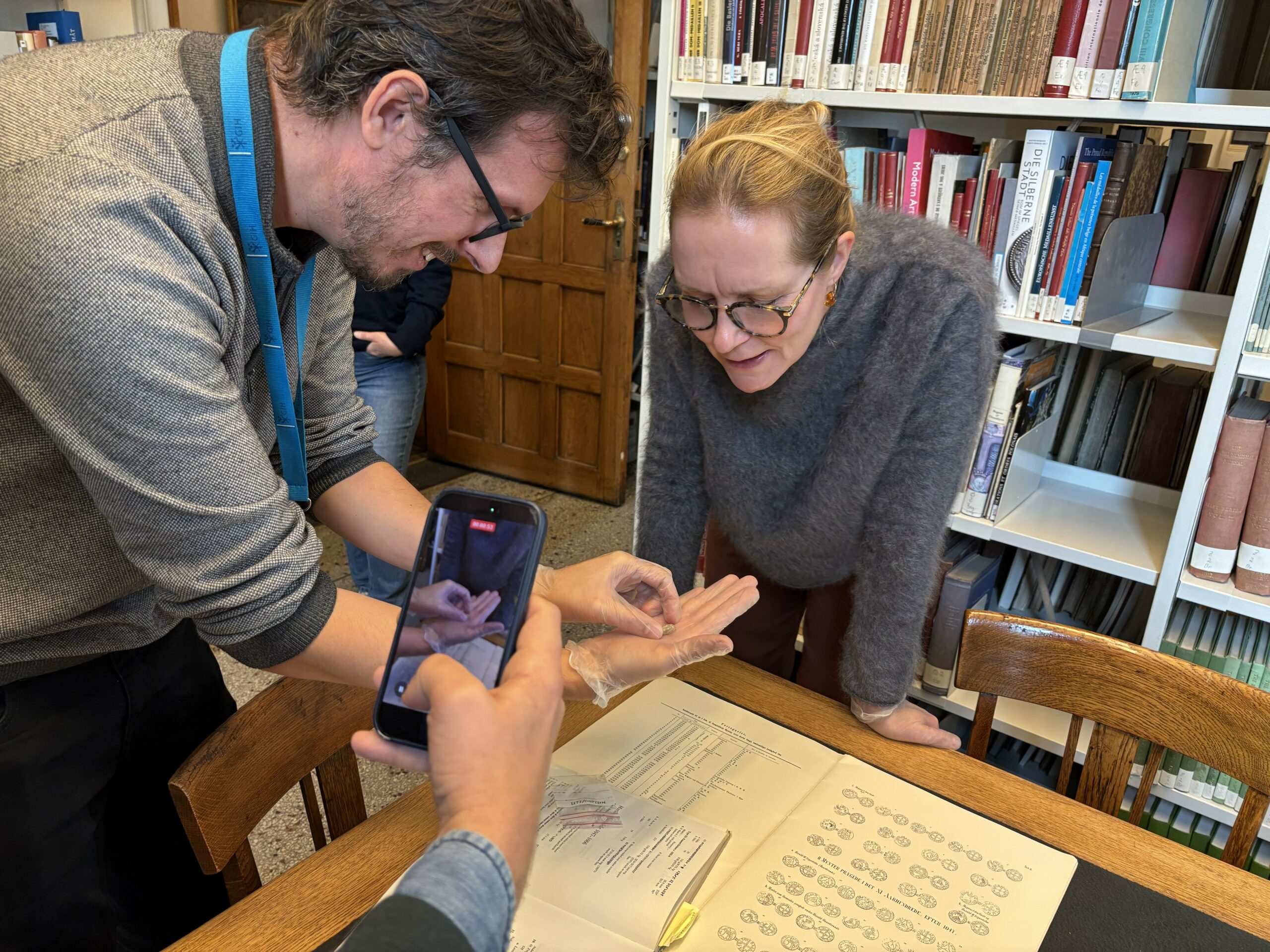 En mann bruker en smarttelefon til å ta bilde av en person som har på seg en plasthanske og holder et lite objekt over en åpen bok med diagrammer.