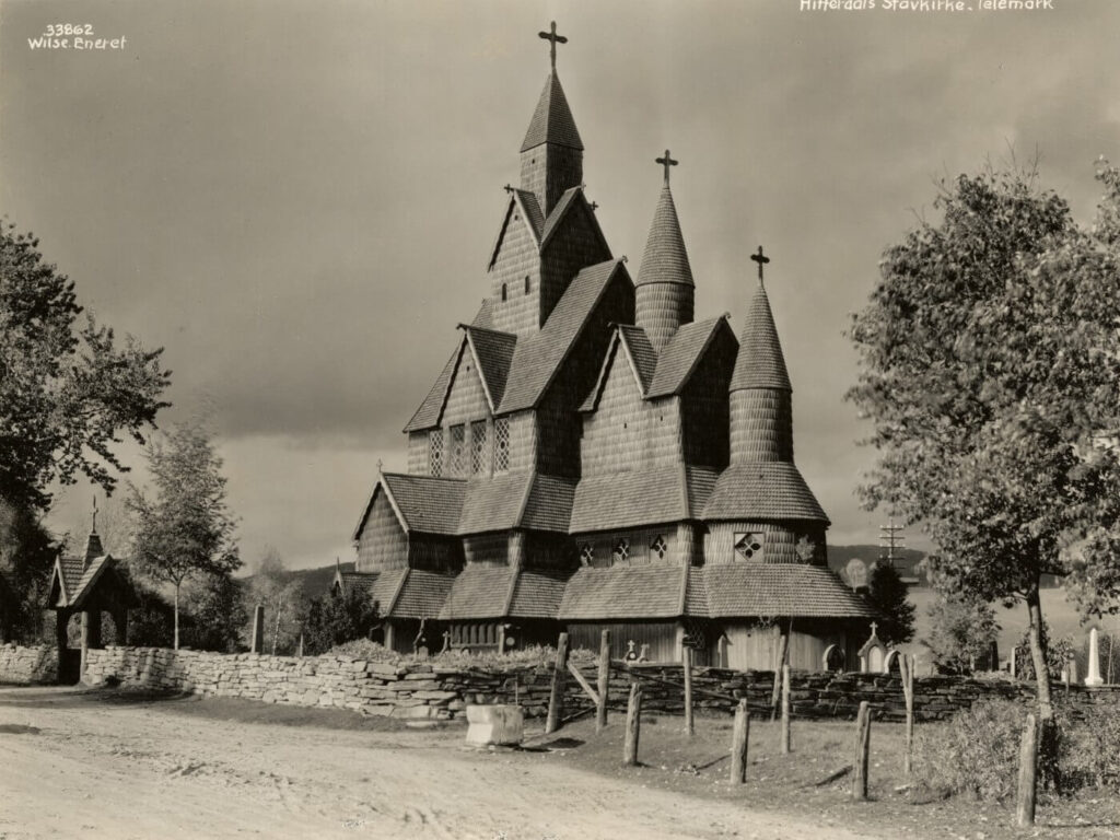 Heddal stavkirke i Telemark, Norge, sett fra en grusvei med en steinmur og et kirkegårdsgjerde i forgrunnen.