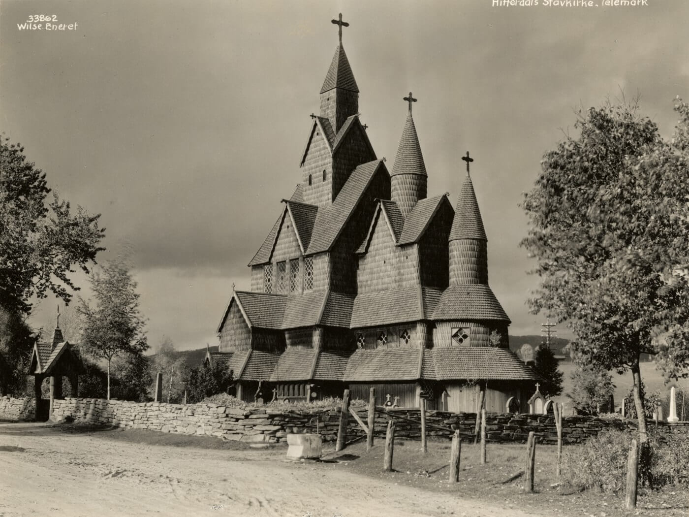 Heddal stavkirke i Telemark, Norge, sett fra en grusvei med en steinmur og et kirkegårdsgjerde i forgrunnen.