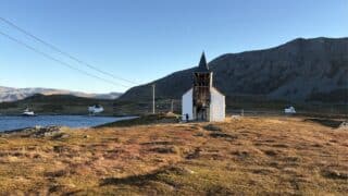 En kirke med en åpen, skjelettlignende fasade som ligger i et kystlandskap med fjell i det fjerne.