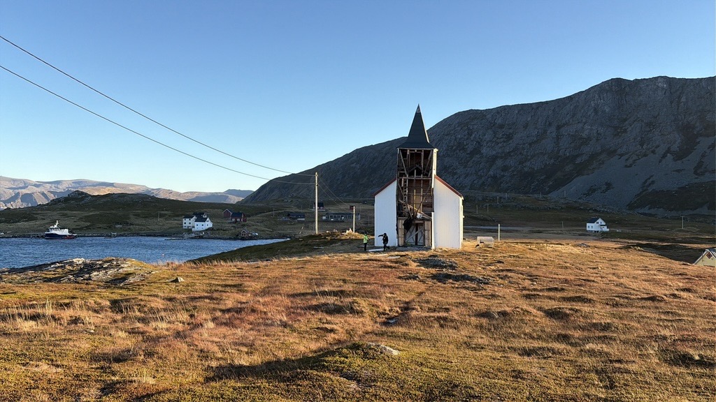 En kirke med en åpen, skjelettlignende fasade som ligger i et kystlandskap med fjell i det fjerne.