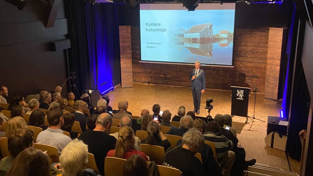 En person i dress står på en scene og snakker til et publikum foran en projeksjonsskjerm med teksten Kystens kulturmiljø – konferanse i Bergen.