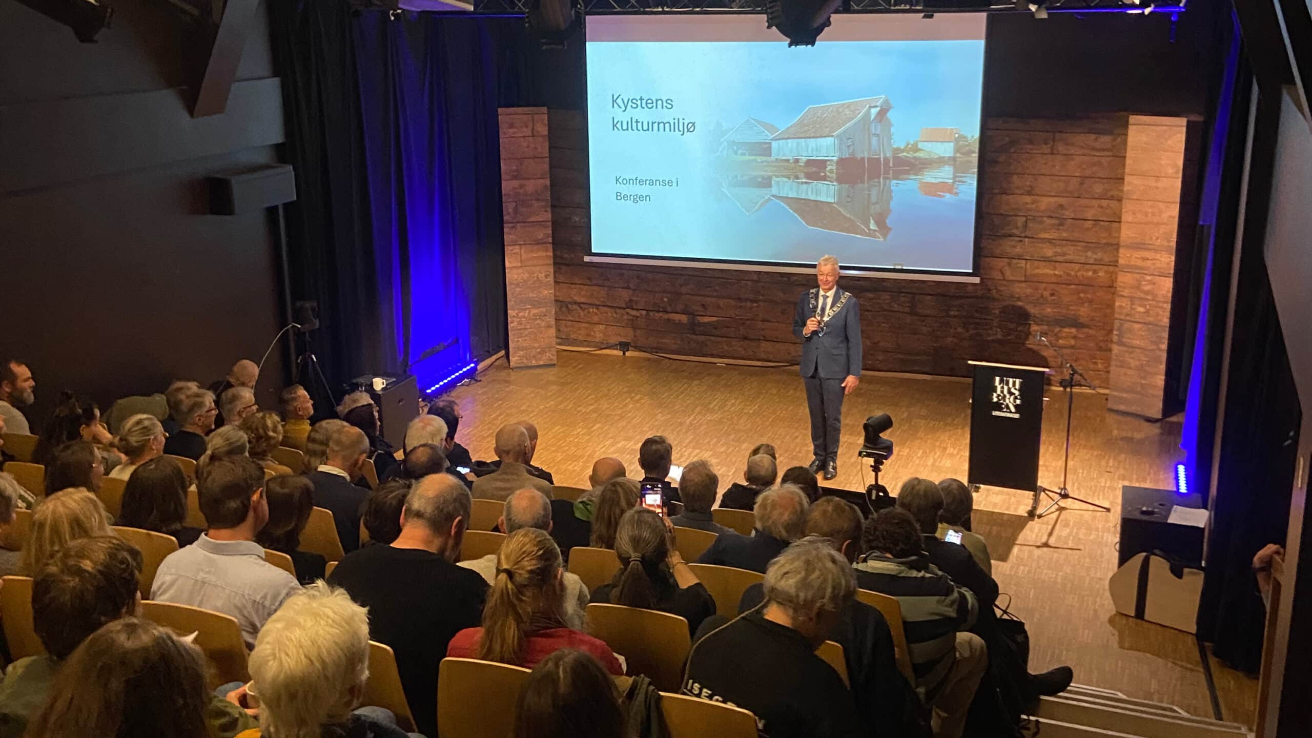 I kø for kystkonferanse 1 En person i dress står på en scene og snakker til et publikum foran en projeksjonsskjerm med teksten Kystens kulturmiljø – konferanse i Bergen.
