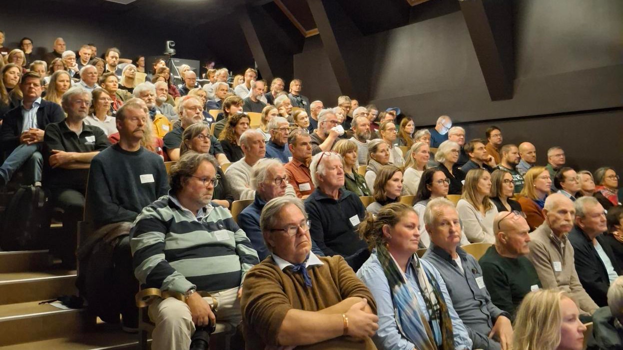 En stor, mangfoldig gruppe voksne som sitter i et auditorium med tribuner og ser på en presentasjon.