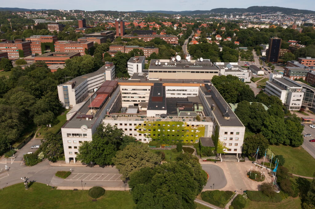 Luftfoto av et stort kontorkompleks med en sentral gårdsplass og en fasade som delvis er dekket av eføy, omgitt av trær og boligbygg.