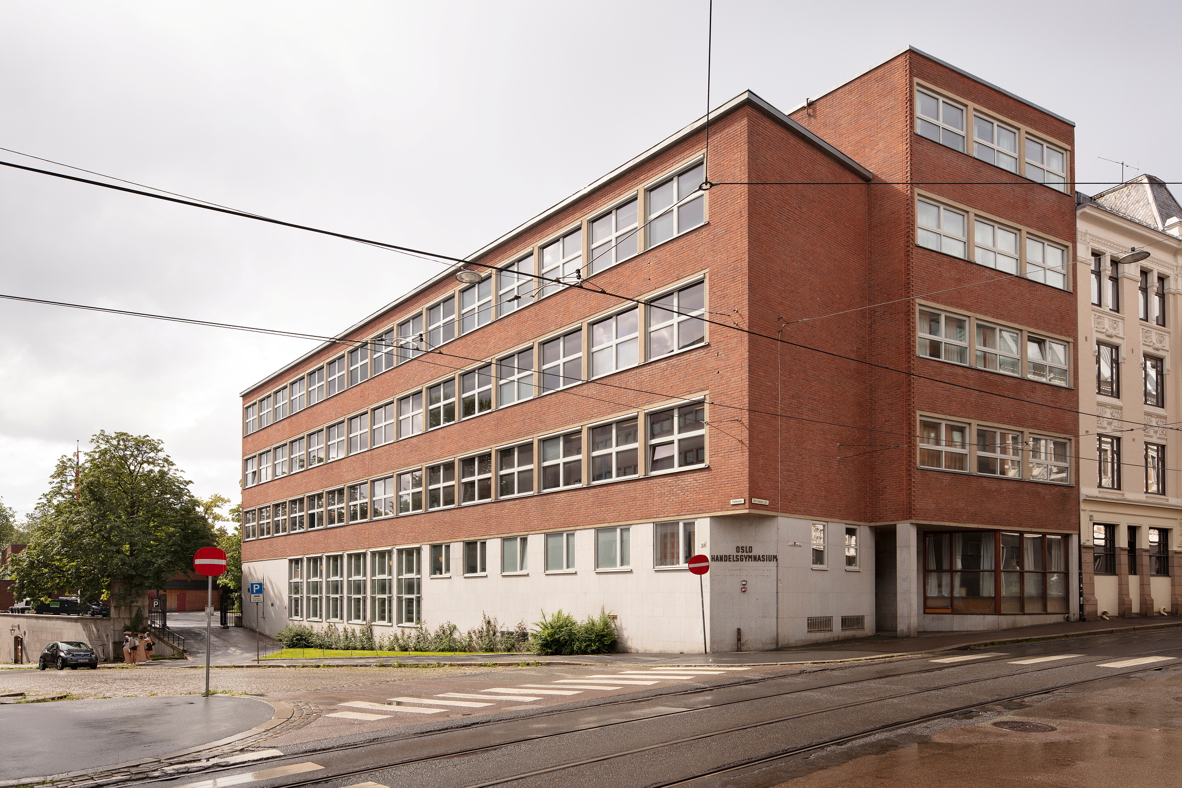 En bygning i rød murstein med skiltet Oslo Handelsgymnasium på et gatehjørne med trikkespor.