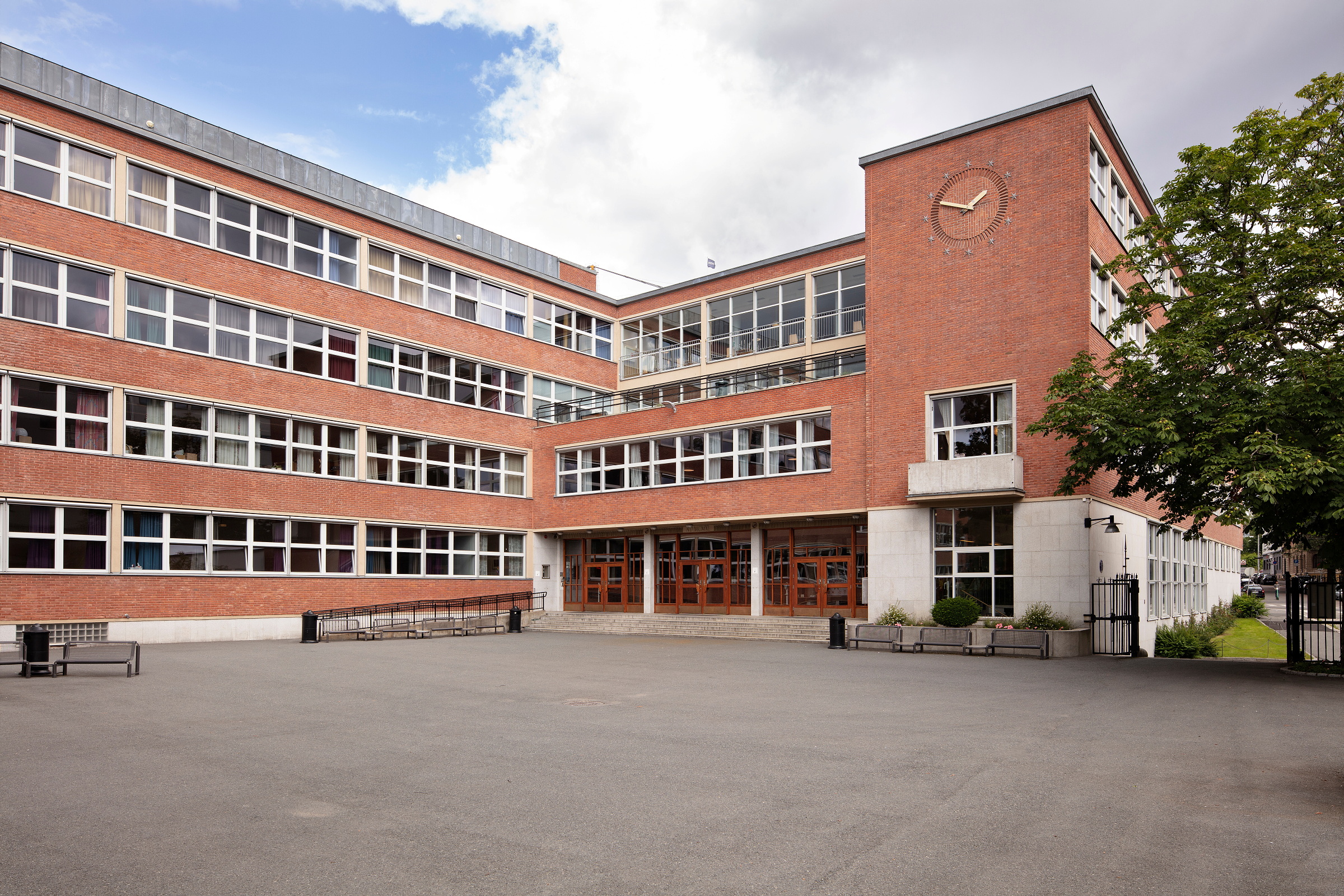 En skolebygning i rød murstein med store vinduer, et klokketårn i hjørnet og en bred, asfaltert gårdsplass ved hovedinngangen.