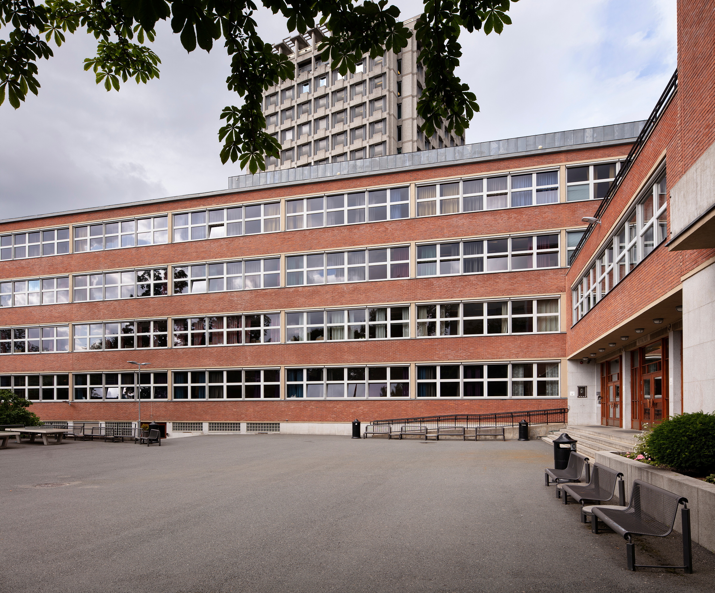 En skolebygning i murstein med flere etasjer, med en stor asfaltert gårdsplass og metallbenker.