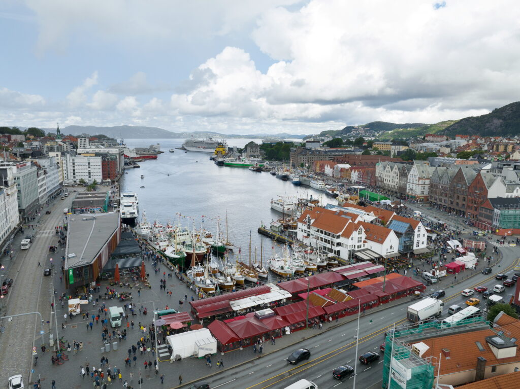 Luftfoto av en travel havn med båter, markedsboder og folkemengder, omgitt av bybygninger, veier og fjell i det fjerne under en delvis overskyet himmel.