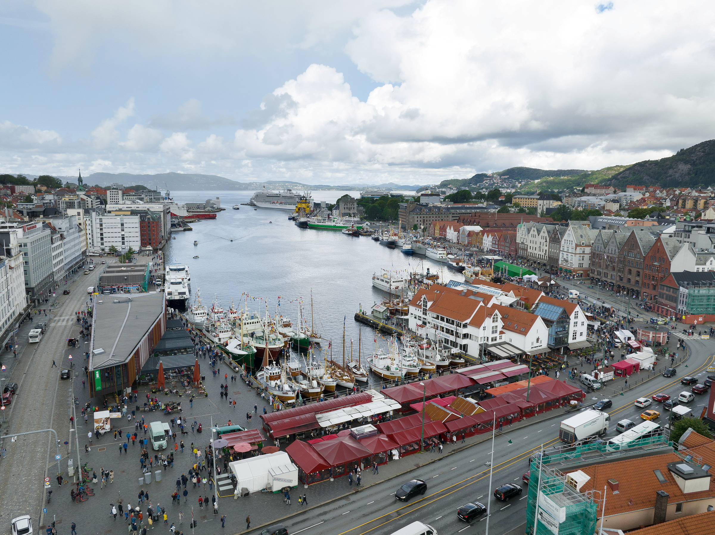Den største undersøkelsen av kulturminner under vann i Norge 1 Luftfoto av en travel havn med båter, markedsboder og folkemengder, omgitt av bybygninger, veier og fjell i det fjerne under en delvis overskyet himmel.