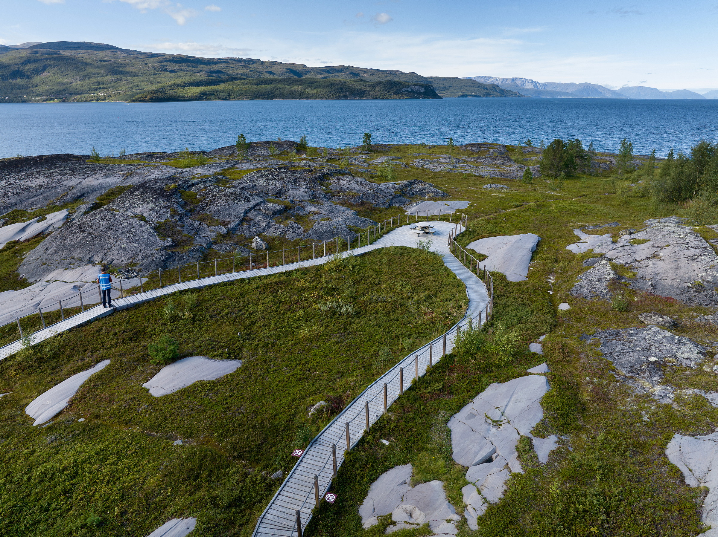 En svingete strandpromenade snor seg gjennom gresskledd terreng og over steiner ved en innsjø, med fjell i bakgrunnen og en person som står ved rekkverket.