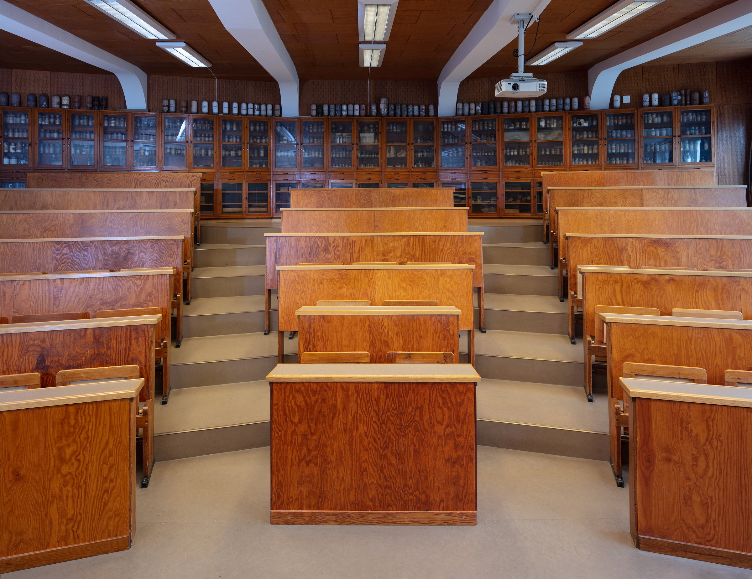 En tom forelesningssal på universitetet med trepulter i trappetrinn og rekker av glasskap fylt med forskjellige glass i bakgrunnen.