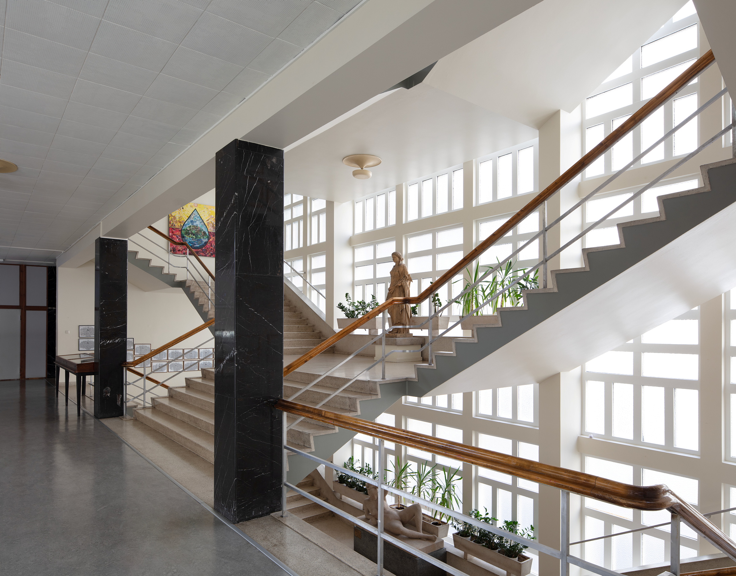 En bred trapp med steintrinn og rekkverk i tre i et solfylt atrium, med en statue av en kvinne og potteplanter.