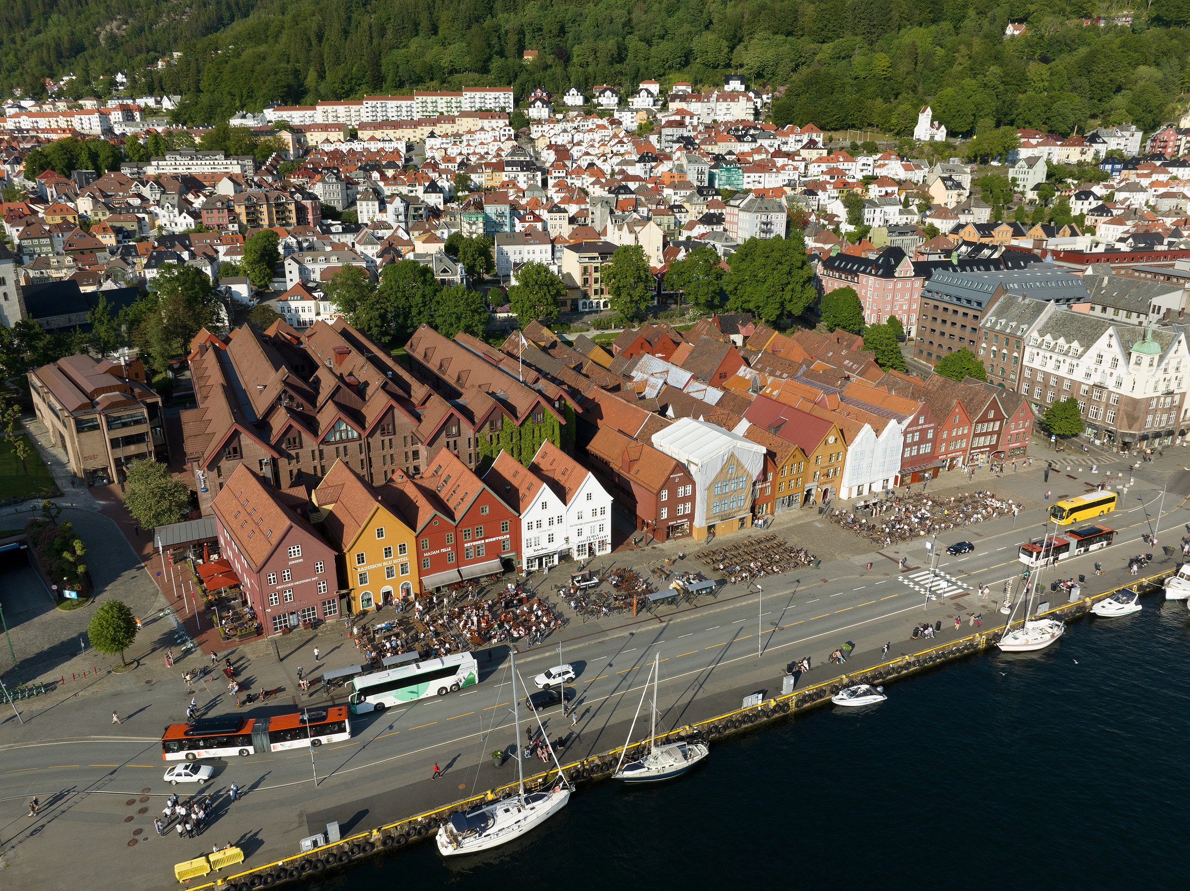 Riksantikvaren reiser ikkje motsegn 1 Luftfoto av fargerike, historiske bygninger langs en gate ved vannkanten med uteservering, busser, biler og båter i havnen, omgitt av et tettbebygget nabolag i åssiden og grønne områder.
