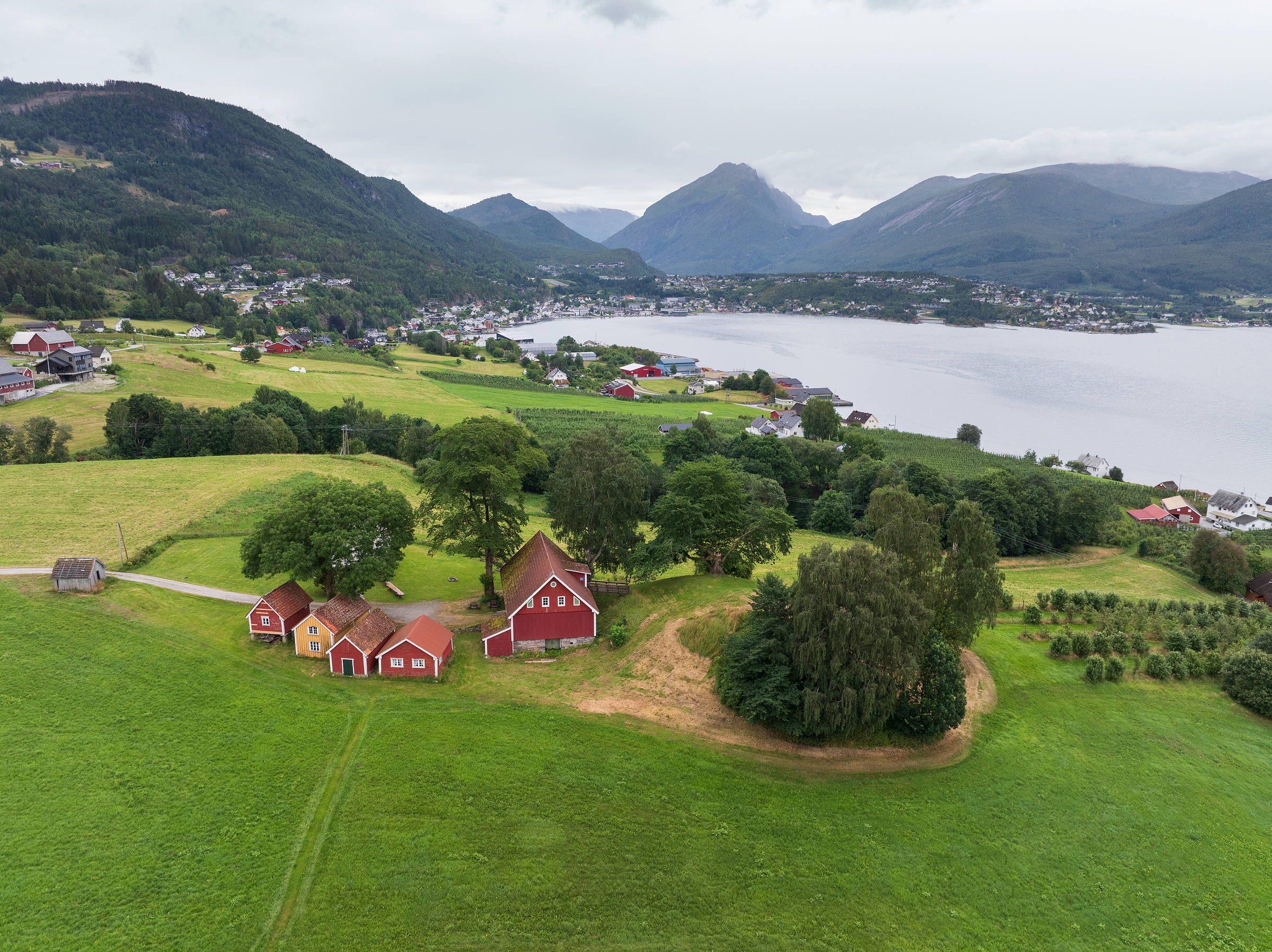 Luftfoto av røde gårdsbygninger på grønne åkrer ved en innsjø, med fjell og en landsby i bakgrunnen under en overskyet himmel.