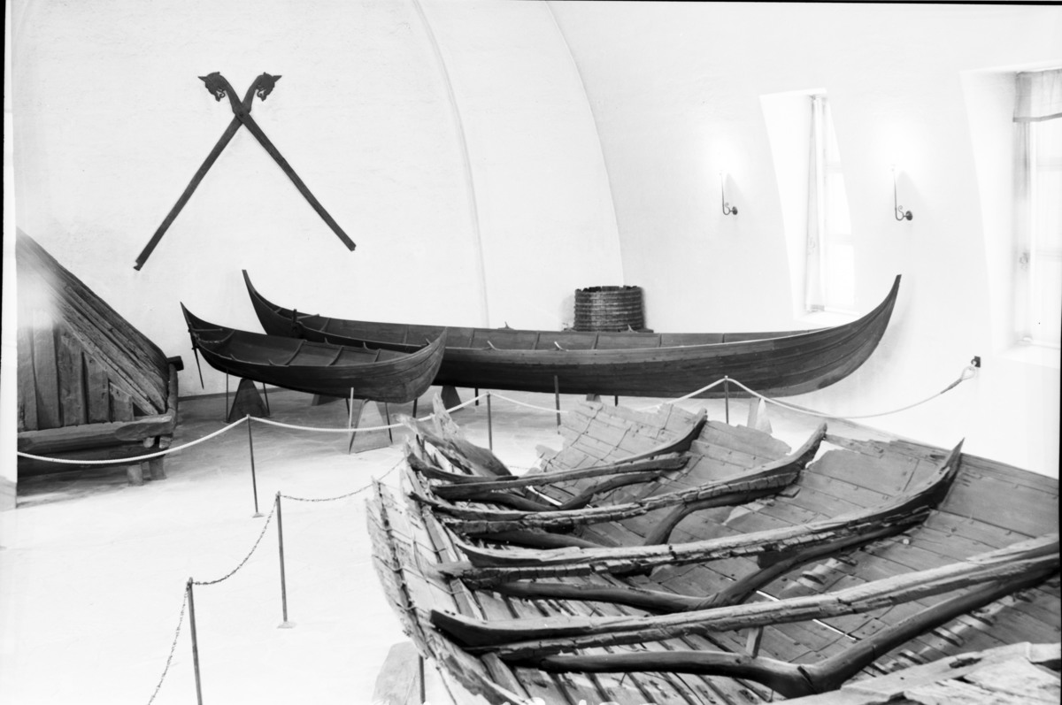 To trebåter i vikingstil utstilt i et museumsgalleri, med det fragmenterte skroget til et større fartøy i forgrunnen.